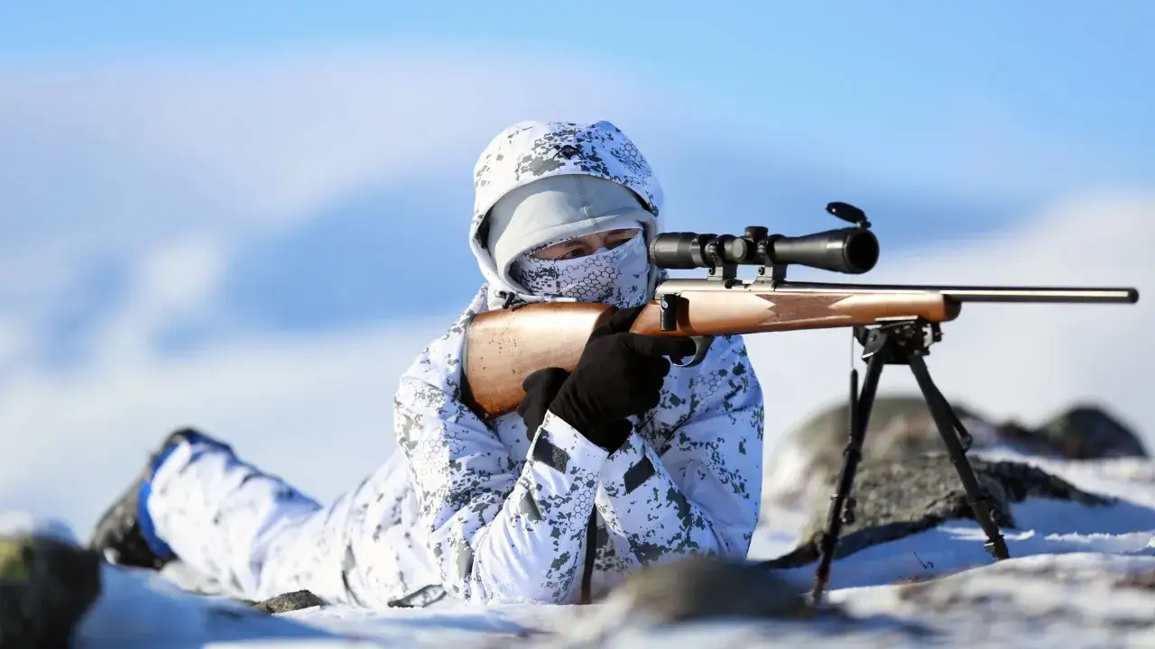 Person i kamouflerad vinterdr&auml;kt ligger i sn&ouml;n och siktar med ett gev&auml;r med kikarsikte. Kanske f&ouml;r att snara ripa i den karga svenska naturen.