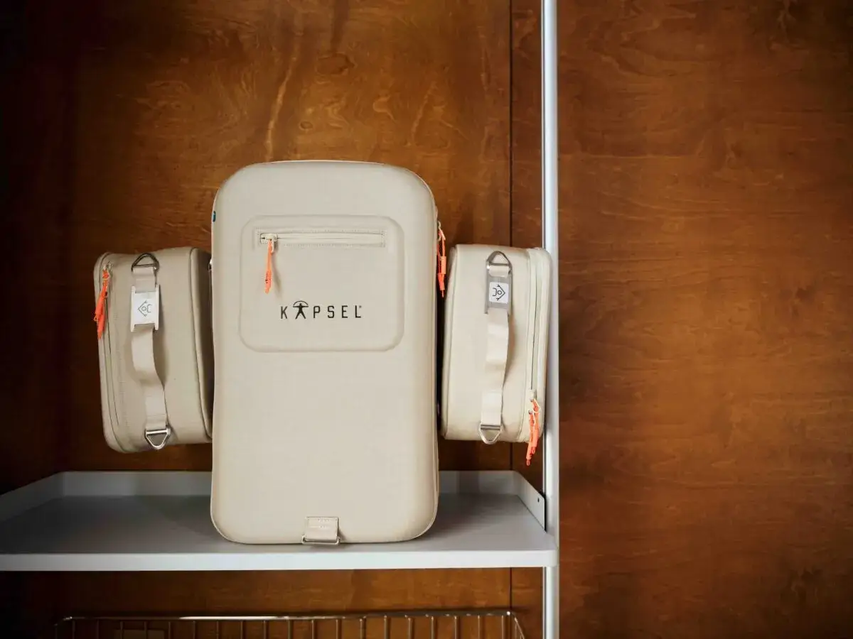 En beige Kapsel bob väska med orange detaljer står på en hylla. Bredvid den hänger två mindre necessärer.