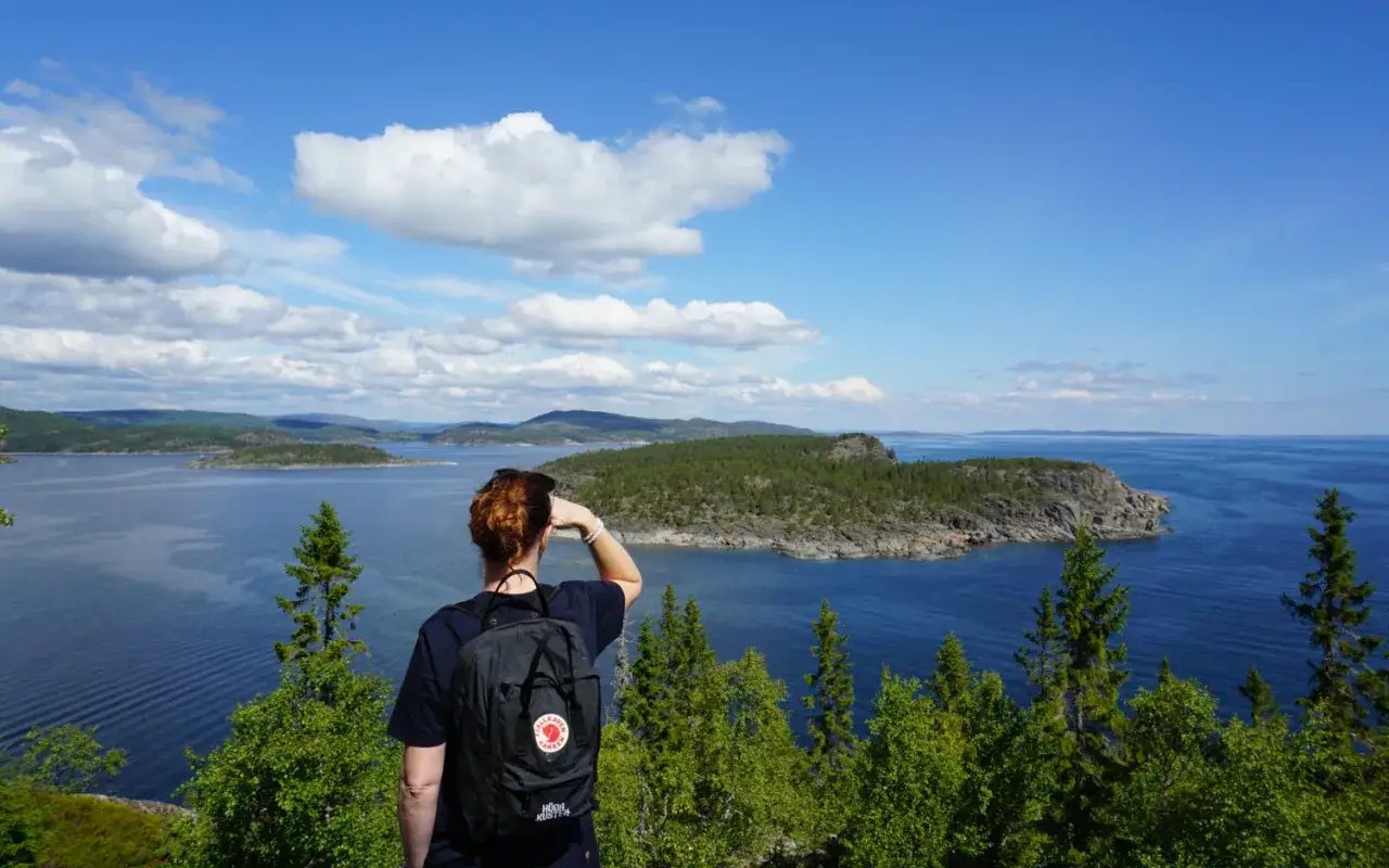 Person med ryggsäck blickar ut över en vidsträckt sjö och skogklädda öar. En vy över Höga Kusten en solig dag.
