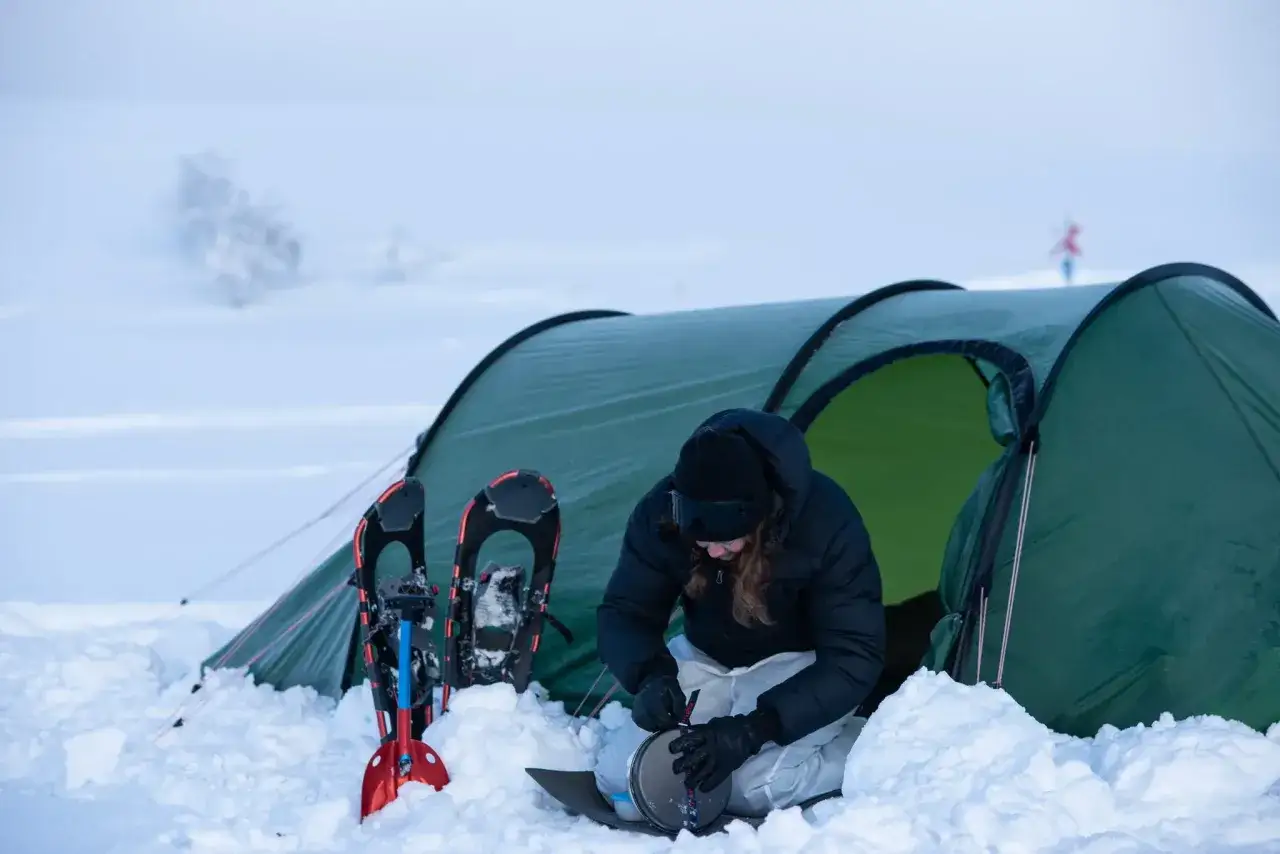 Person förbereder mat vid ett tält i snön, med snöskor och spade redo. Snart blir det värme i bilen på vintern.