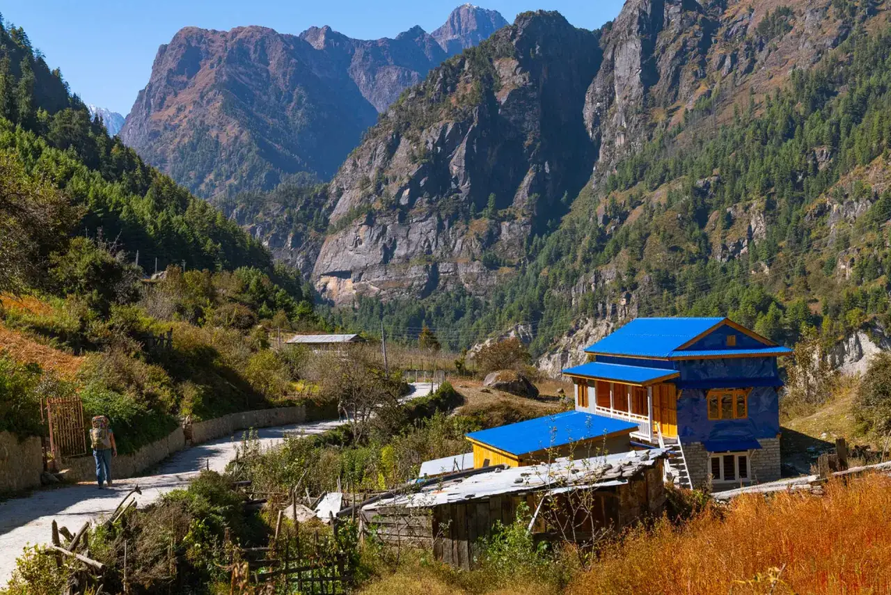 En person vandrar i Nepal längs en grusväg i en dal omgiven av höga berg och gröna skogar. Ett hus med blå tak syns i förgrunden.