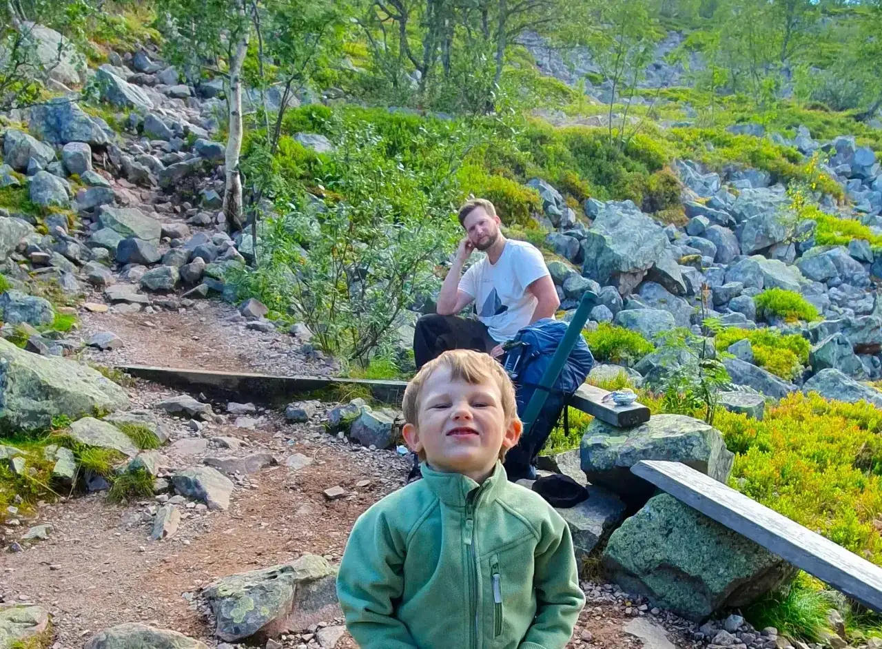 Glad pojke i grönt klär sig för att vandra med barn i fjällen. Pappa vilar bakom.