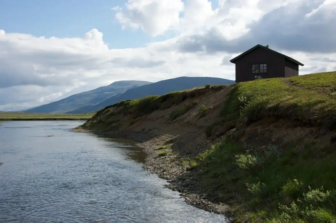 En mörk stuga på en grön kulle vid en forsande älv i Padjelanta nationalpark. Berg i bakgrunden under en molnig himmel.