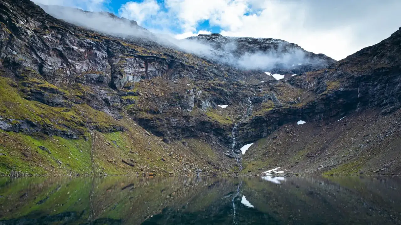En spegelblank sjö reflekterar en dramatisk bergssida med en forsande vattenfall nära Abisko Turiststation. Moln sveper över topparna.