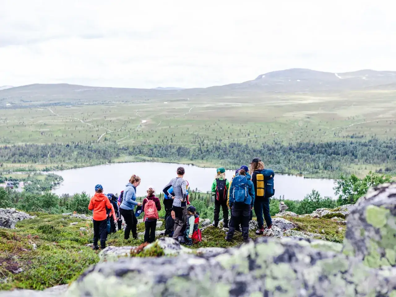 En grupp människor, inklusive barn, deltar i guidade vandringar i ett vackert fjällandskap med en sjö i bakgrunden.