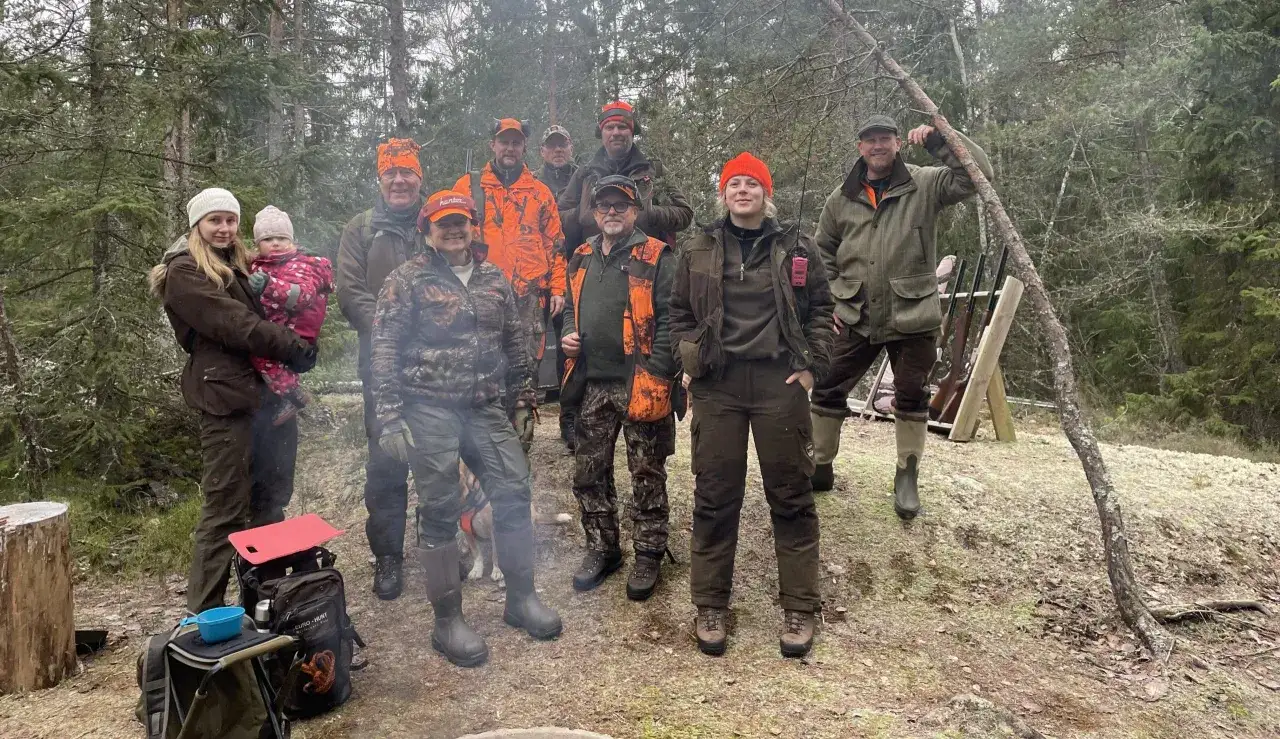 En grupp jägare i skogen, inklusive en kvinna med barn. De är klädda i jaktkläder. Ingen indikation på jaktbrott eller straff.