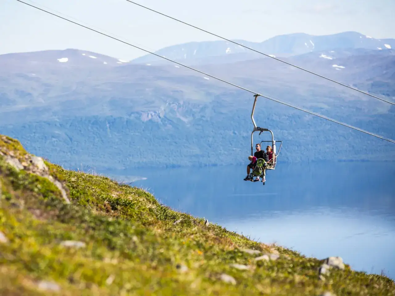 Två personer och ett barn åker linbana över en sjö med berg i bakgrunden. En vy som påminner om en aurora sky station.