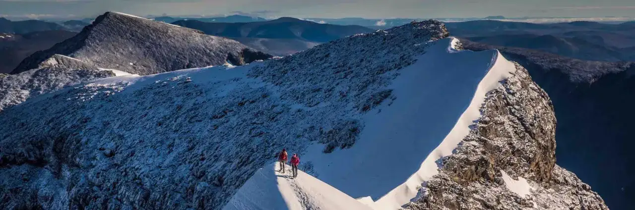 Två vandrare på en snöig bergskam, nära Sveriges högsta berg. Dramatiskt landskap med mörka berg i bakgrunden.