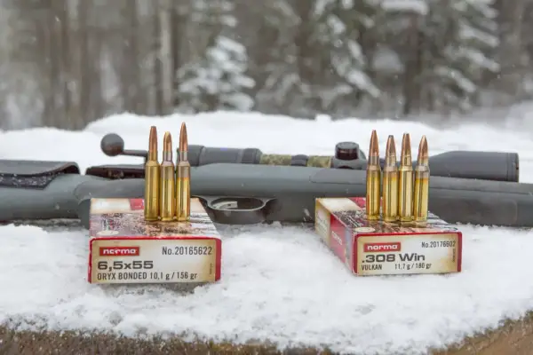 Två lådor med Norma ammunition, 6,5x55 och .308 Win, står på snö framför ett gevär. Klass 1 ammunition redo för jakt.