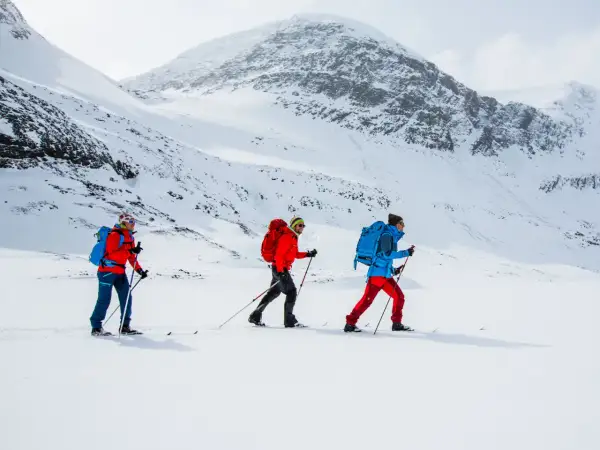 Tre personer åker skidor i ett snöigt landskap nära Abisko. De är klädda i färgglada jackor och ryggsäckar, redo för äventyr.