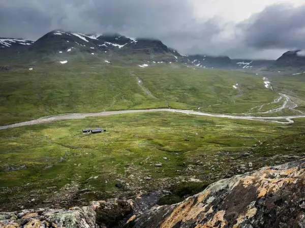 Grön dal med en flod och fjällstugor, som en karta över Kungsleden. Moln hänger över snöklädda berg.
