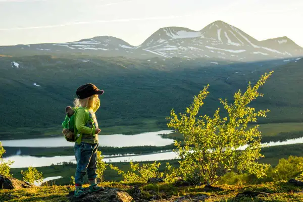 Ett barn med ryggsäck blickar ut över en vidsträckt fjällvy med sjöar och snöklädda toppar. En perfekt bild av vandring med boende i naturen.