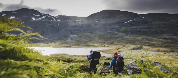 Två vandrare med ryggsäckar går mot en sjö i fjällen. En karta över midnattssolens utbredning kan hjälpa dem att planera sin tur.